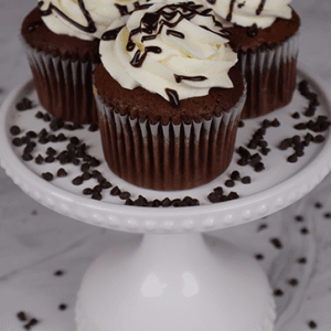 Three CC-006 Display Chocolate whipped cream Cupcakes with swirled white frosting and chocolate drizzle are on a white cake stand, surrounded by chocolate chips.