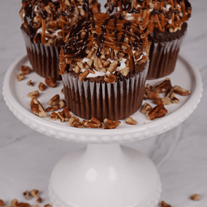Three CC-000V Turtle Cupcakes sit on a white cake stand, each topped with caramel, chopped pecans, and chocolate drizzle, with scattered pecan pieces around them.