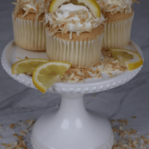 Three CC-000X Lemon Cupcakes topped with whipped cream, toasted coconut flakes, and lemon slices are displayed on a white cake stand, with extra lemon slices and coconut flakes around them.