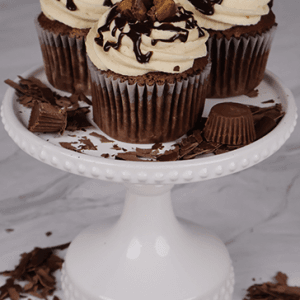 Three CC-000S Chocolate Peanut Butter cupcakes with swirled frosting, chocolate drizzle, and peanut butter cup pieces are displayed on a white cake stand, surrounded by extra chocolate and peanut butter cup candies.