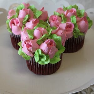 Three CC-086 chocolate cupcakes with white filling, decorated with pink and green frosting to look like blooming roses and leaves, arranged on a white cake stand.