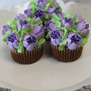 Three CC-088 Chocolate cupcakes with white filling, decorated with purple and green buttercream flowers, are displayed on a white scalloped cake stand.