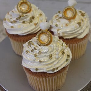 Three CC-093 chocolate cupcakes with white swirled frosting, gold and pearl sprinkles, each topped with an edible engagement ring and diamond, displayed on a silver stand.