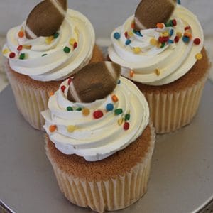 A gold cupcake with chocolate filling, topped with swirled white frosting, colorful round sprinkles, and a small chocolate football decoration, displayed on a silver cake stand.