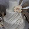 The WD-020 is a two-tier white cake with pleated fondant, beige sugar flowers, and dried grass accents, presented on a silver cake board against a wooden background.