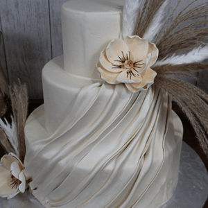 The WD-020 is a two-tier white cake with pleated fondant, beige sugar flowers, and dried grass accents, presented on a silver cake board against a wooden background.