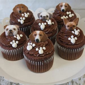 Six CC-099 chocolate cupcakes with white filling, decorated with fondant dog faces and small white paw prints, arranged on a white cake stand.