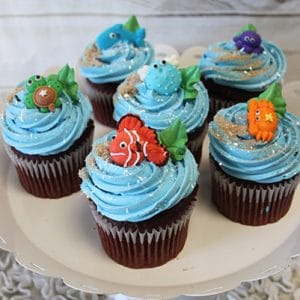 Six CC-105 chocolate cupcakes with white filling are topped with edible sea creature decorations, including colorful fish and turtles, and displayed on a white cake stand.