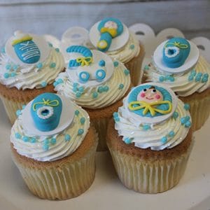 Six CC-109 chocolate cupcakes with white filling, topped with blue, yellow, and white baby-themed fondant decorations like bottles, rattles, diaper pins, and a baby's face, displayed on a white stand.