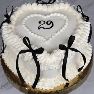 BD-023e Heart shape one size: White frosted heart-shaped cake with piped icing, black ribbon bows on the sides, and the number 29 inside a heart outline on top. Presented on a gold board placed on marble.