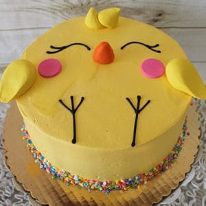 EA-028 is a round yellow cake designed as a cute chick, featuring fondant wings, beak, cheeks, closed eyes, feet, and colorful sprinkles at the base on a gold cake board.