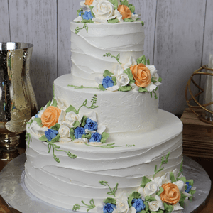 The WC-000J is a three-tiered white cake adorned with orange, blue, and white buttercream flowers plus green leaves, set on a silver base against a light wood background.