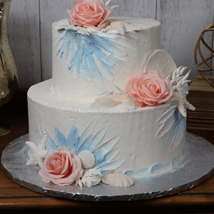 The WD-038 is a two-tiered white cake with blue painted accents, large pink icing roses, and white fondant leaves and seashells, displayed on a wooden table.