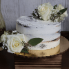 A rustic, semi-naked BR-005 Chocolate cake/chocolate mousse decorated with white roses, greenery, and baby's breath, presented on a gold cake board atop a wooden box against a wooden backdrop.
