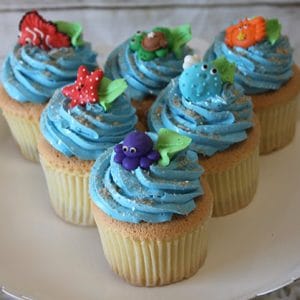 Six CC-106 Gold cupcakes with chocolate filling are topped with blue frosting and fondant sea creatures—fish, starfish, turtle, pufferfish, octopus, and crab—displayed on a white cake stand.