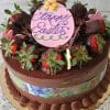 A Tiramisu Cake decorated with chocolate-dipped strawberries, chocolate shavings, and a pink oval topper reading Happy Easter in purple icing sits on a gold base with a festive Easter-themed band around the side.