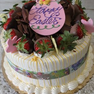 A Mango Cake decorated with chocolate-covered strawberries, chocolate curls, and pink candy bunnies, topped with a pink "Happy Easter" plaque and wrapped in a festive Easter-themed ribbon.