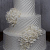 The WC-001B is a three-tiered white wedding cake with textured diagonal lines, adorned with clusters of white buttercream roses. Displayed on a silver board, it stands out beautifully against a wooden background.