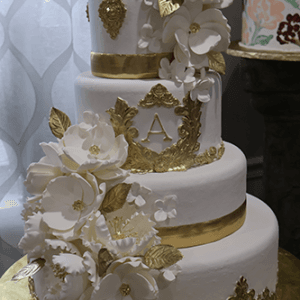 WD-001B is a three-tiered white cake adorned with gold accents, ornate trim, large white sugar flowers, and a gold monogram A on the middle tier, displayed on a gold-covered base.
