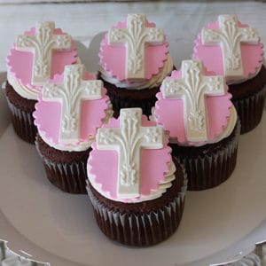 Six CC-040 Chocolate cupcakes with white chocolate mousse filling, topped with pink and white icing and a white ornate cross on a pink fondant circle, displayed on a white plate.