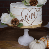 The BR-000F is a white frosted cake featuring white roses, pinecones, and greenery on a white stand. An icing heart with K+M and arrow decorates the front. Two small pumpkins are displayed next to the stand.