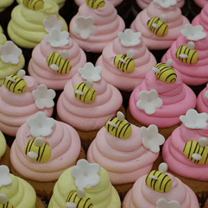 Rows of CC-139 Chocolate cupcakes with white filling, topped with pastel pink and yellow frosting, each adorned with fondant bees and white fondant flowers.
