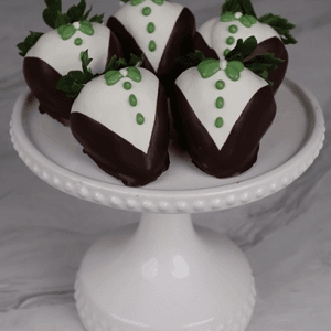 DB-000G Groom Strawberries: Five strawberries dressed in dark and white chocolate tuxedos with green icing details, presented on a white cake stand against a marble background.