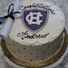 The GC-000S Holy Cross is a round cake with white icing, featuring a purple and white shield emblem, “Congratulations Andrew,” and a diploma tied with a black ribbon, displayed on a white rose-patterned tablecloth.