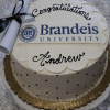 A round GC-000P Brandeis University cake with white frosting, black dots, a gold base, "Congratulations Andrew," the Brandeis logo, and a diploma decoration with a black ribbon on top.