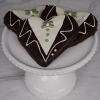 Three DB-000L Groom Cookies shaped like hearts and decorated with white, brown, and green icing to resemble tuxedos are arranged on a white pedestal cake stand against a marble background.