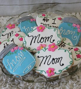 A plate of CE-050d Mother's Day cookies shaped like jars and plaques, decorated with “Mom,” “#1 Mom,” pink flowers, and blue or white icing in cursive.