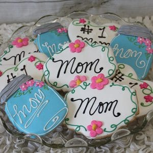 A plate of CE-050d Mother's Day cookies shaped like jars and plaques, decorated with “Mom,” “#1 Mom,” pink flowers, and blue or white icing in cursive.
