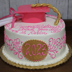 A round graduation cake (GC-019) with pink and gold icing, 2023 on the side, a pink grad cap topper, and “Congratulations Grad, You did it Robin” written on it.