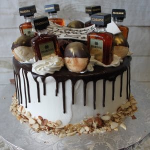 The BD-024E Disaronno is a round cake with white frosting, chocolate drip, small Disaronno bottles on top, gold truffles, cream swirls, and a base edged with sliced almonds.