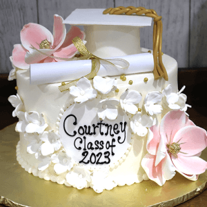 GC-044 is a white graduation cake adorned with pink and white flowers, a fondant diploma, and a graduation cap, featuring “Courtney Class of 2023” on a white plaque.