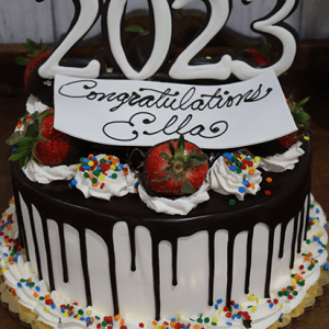 GC-011 is a chocolate-dripped cake topped with whipped cream, strawberries, rainbow sprinkles, and a "2023" topper. The cake features a cursive sign reading “Congratulations Ella.”.