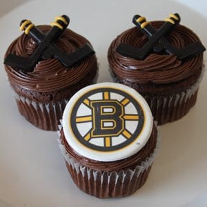 Three CC-117A chocolate cupcakes with white filling, topped with chocolate frosting. Two feature hockey stick decorations; the third displays a white fondant circle with the Boston Bruins logo.
