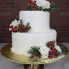 The TC-000N is a two-tiered white cake adorned with snowflakes, pinecones, red berries, white roses, and green foliage, presented on a gold stand against a brick wall backdrop.