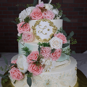 The WD-001 is a two-tiered white cake with pink, white, and cream flowers, gold accents, an ornate L monogram, a gold "15" topper, and sits on a gold board.