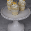 Three small CO-104 Mango Shooters of layered yellow and white mousse, topped with whipped cream and yellow chocolate decorations, displayed on a white beaded-edge cake stand against a marble background.