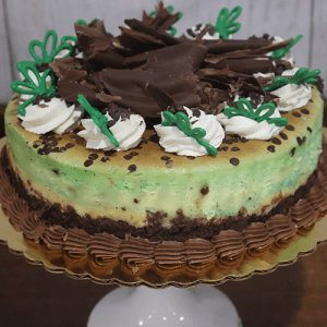 The GD-016 Mint Chocolate Chip Cheesecake features round green and brown layers, topped with whipped cream, chocolate shavings, green icing accents, and chocolate chips on a gold cake board and white stand.