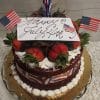 The JC-002 Red Velvet is a festive cake adorned with strawberries, chocolate pieces, mini American flags, a “Happy July 4th” card, and red, white, and blue accents. It sits on a gold tray for a celebratory touch.