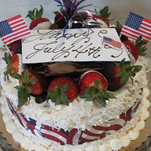A festive JC-007 Pina Colada cake adorned with strawberries, chocolate, small American flags, red, white, and blue ribbon, plus a 'Happy July 4th' card in cursive sits on a glass platter.