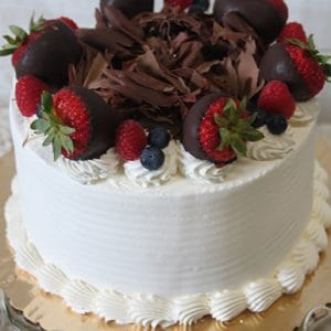 The BD-018D Triple Berry Cake features a white frosted finish, chocolate shavings, chocolate-covered strawberries, raspberries, and blueberries on a scalloped gold cake board.
