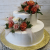 WD-007: A two-tier white cake adorned with pink, red, and white roses, greenery, and delicate sprigs, presented on a gold cake board against a light wood background.