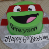 CB-005F is a round green cake decorated as a Ninja Turtle with a red mask, smiling mouth with tongue out, and "Greyson" written on it. A sign in front says "Happy 6th Birthday.