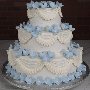 The WD-000D is a three-tiered white cake with piped white frosting swags and clusters of light blue fondant flowers, displayed on a silver cake board against a dark brick background.