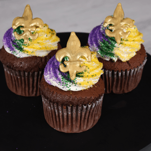 Three CC-142 Chocolate cupcakes with white chocolate mousse filling, swirled purple, yellow, and white frosting, green and gold sprinkles, and a gold fleur-de-lis decoration are arranged on a black plate.
