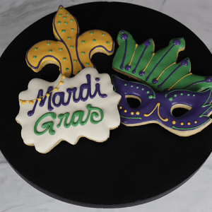 CE-007 Mardi Gras features three decorated cookies on a black plate: a yellow and green fleur-de-lis, a green and purple mask, and a white cookie with “Mardi Gras” in purple and green icing.