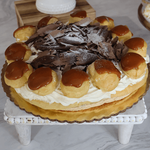 The GD-028 Gateau St. Honore with Caramel features a round pastry topped with caramel-coated cream puffs, filled with whipped cream, and finished with chocolate shavings in the center, presented on a white decorative stand.